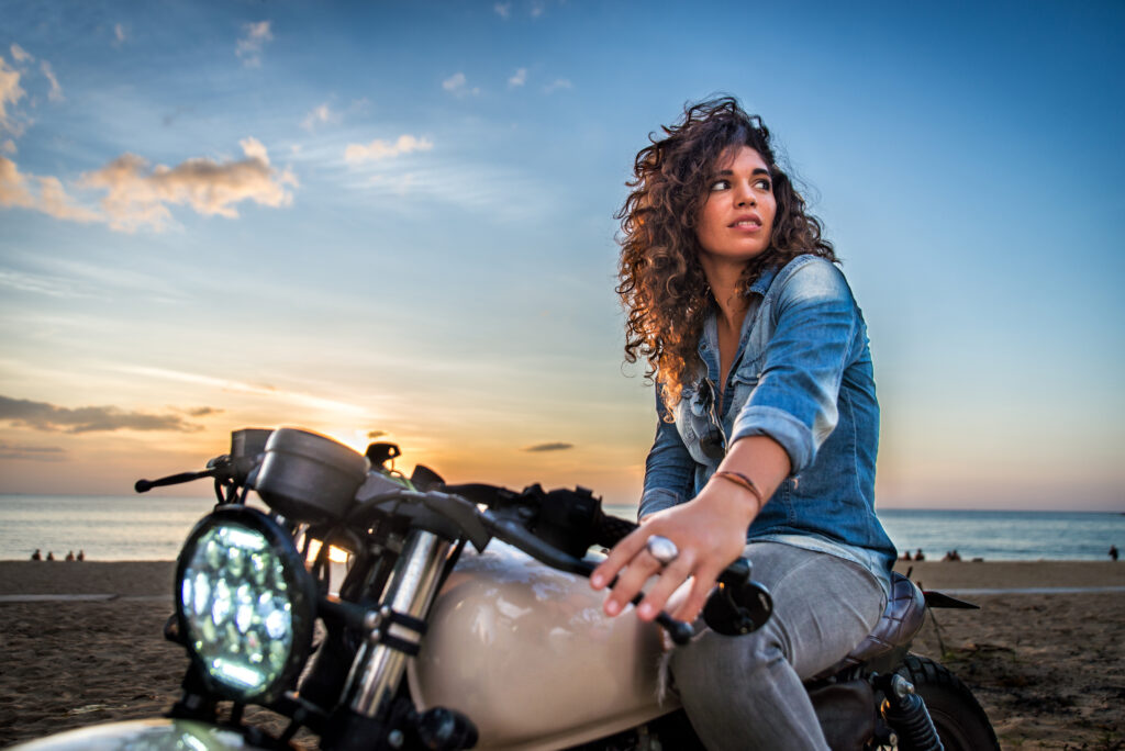Woman sitting on a motorcycle near the beach at sunset, representing motorcycle riding and lifestyle.